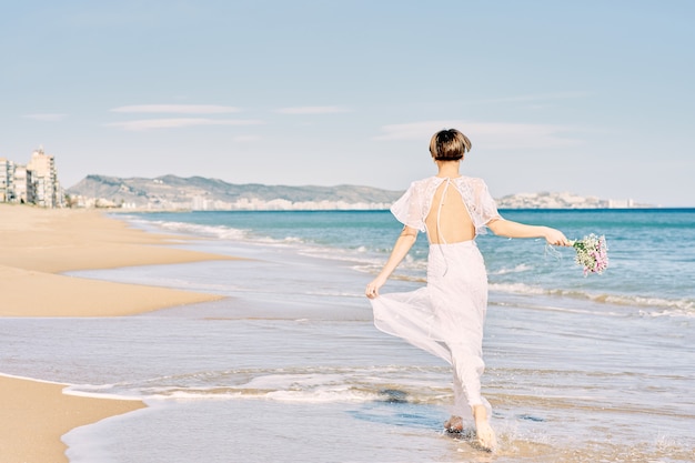 Невеста счастливо бежит по пляжу в свадебном платье и держит в руке букет цветов