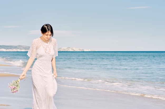 Foto sposa che corre felicemente lungo la spiaggia nel suo abito da sposa e con in mano un mazzo di fiori