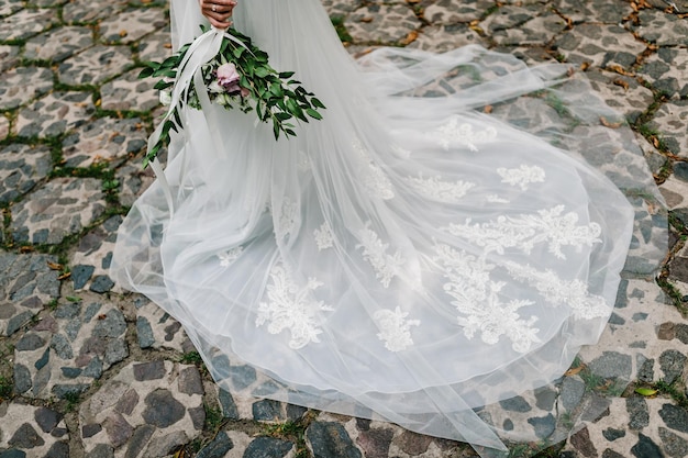 The bride's hand holds from below a wedding bouquet a look at the bottom of the dress and women's feet The girl goes on a stone pavement train to the dress