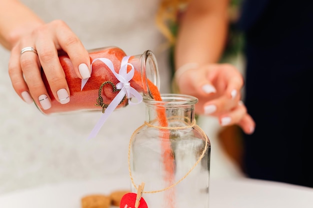 赤い砂の瓶を保持し、それを統一花瓶に注ぐ結婚指輪を持つ花嫁の手