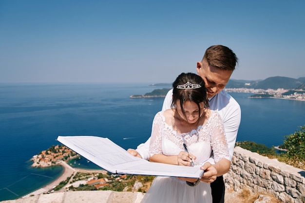 Foto la sposa segna nel libro di registrazione nelle mani dello sposo in piedi sulla montagna sveti stefan