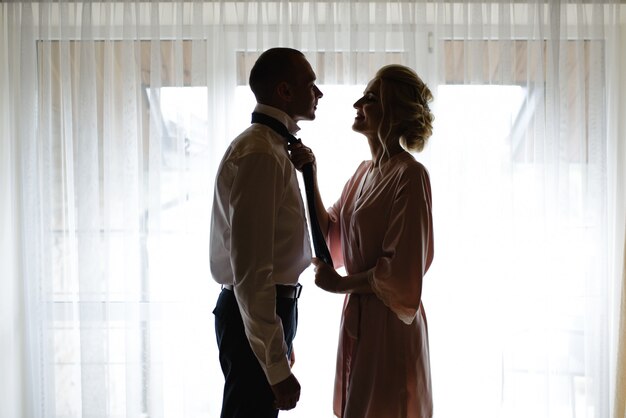 A bride in a silk robe helps put a tie on the groom. The bride and groom are preparing for the wedding together