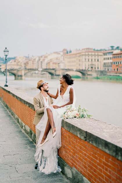 Bride sits on a brick fence next to the groom on the river bank