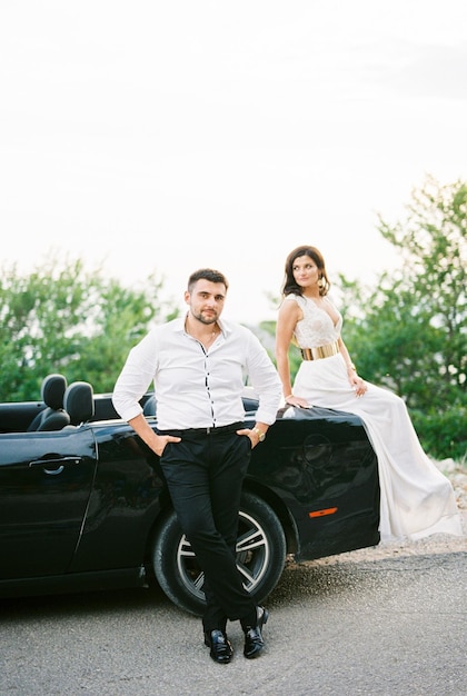 Bride sits on the bumper of a sports convertible next to the standing groom