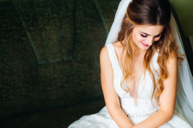 Bride sits on sofa in wedding dress with wedding veil and smiles