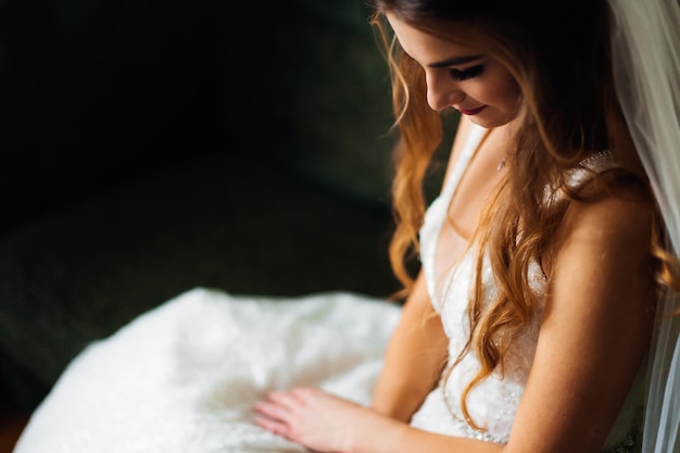 Невеста сидит на диване в свадебном платье с вуалью и улыбается