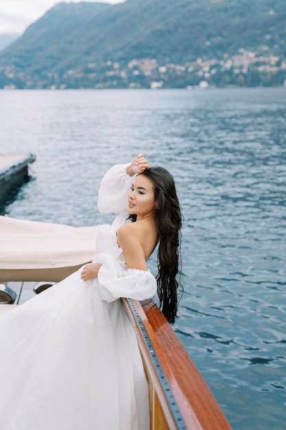 Bride stands leaning on the side of a yacht touching her forehead with her hand floating on the sea