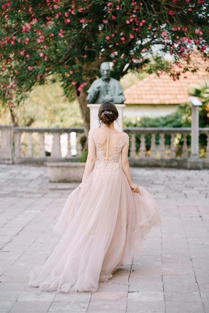 Невеста стоит возле скульптуры под цветущим деревом, вид сзади