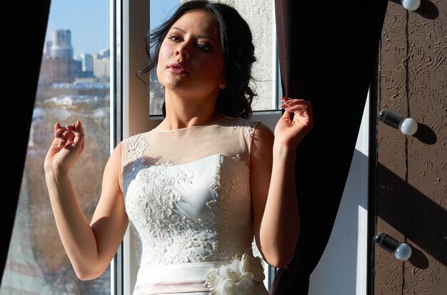 The bride stands near the window and sun ray wedding gown