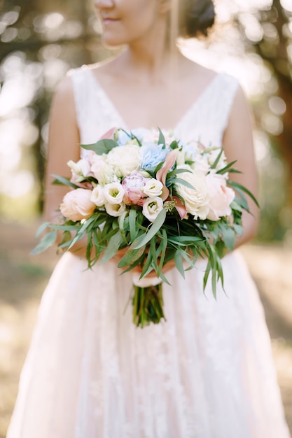 A bride stands in an olive grove and holds in her hands a bouquet with roses peonies lisianthus and