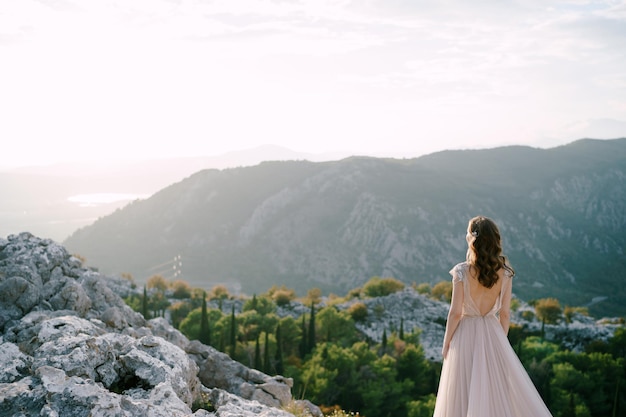 Невеста стоит на скалистой горе и смотрит на залив сзади