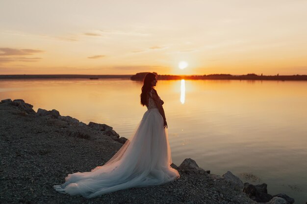 A bride stands on the shore of a lake at sunset.