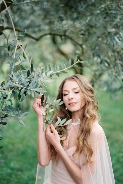 Bride touches with her hands the branches of a green olive tree in a grove