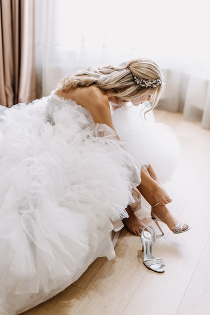 Bride wearing a long white tulle dress, putting on her shoes in the morning.