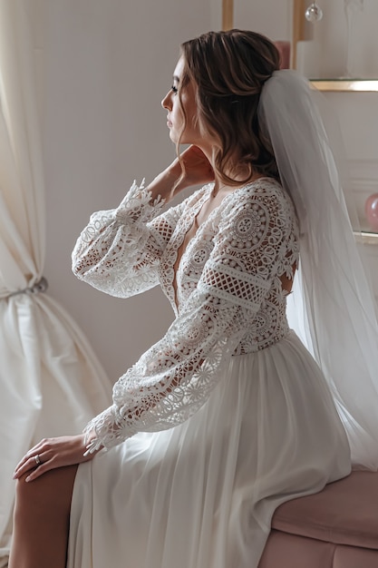 Bride in a wedding dress in boho style and with a veil posing sitting in a cozy room. Wedding photo session, bride's morning.