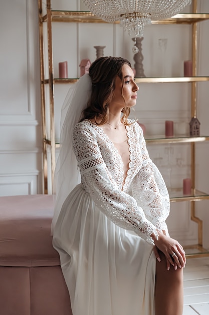 Bride in a wedding dress in boho style and with a veil posing sitting in a cozy room.