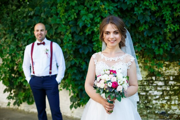 手前に白いドレスを着た花嫁とサスペンダーを持った新郎が後ろに立っています。緑の葉と背景の壁に