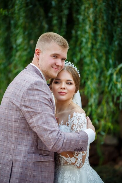 Bride in white dress and groom in costume cuddle and walk in the park