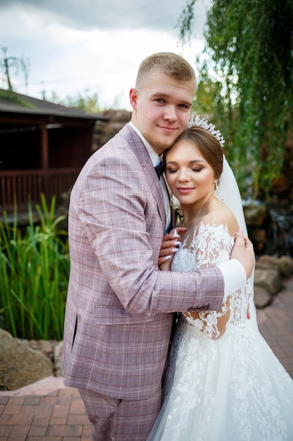 Bride in white dress and groom in costume cuddle and walk in the park