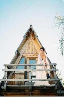 La sposa in abito bianco si trova sul balcone di una casa triangolare appoggiata a una ringhiera di legno