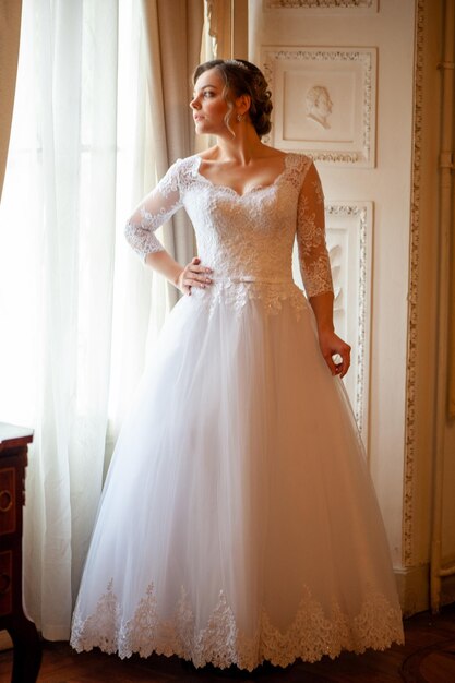 A bride in a white dress stands in front of a window.