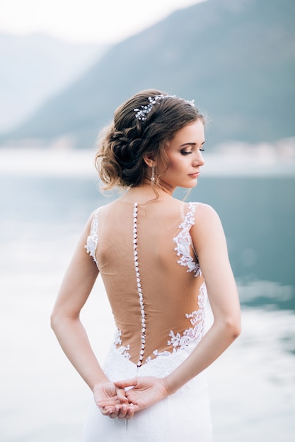 白いドレスを着た花嫁が立って、手を後ろに抱き、海と山を背景に頭を向けます。高品質の写真
