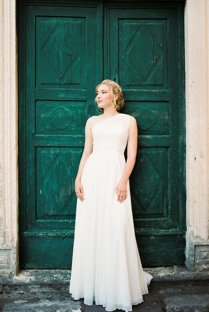Bride in a white dress stands leaning against a green door