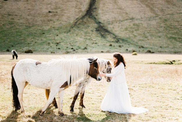 白いドレスを着た花嫁がアイスランドの山のふもとで馬をなでる