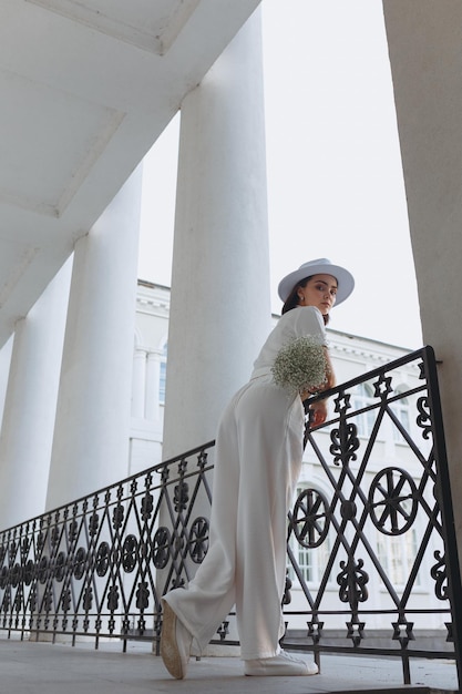 Bride in white outfit and hat standing on balcony with wedding bouquet of flowers