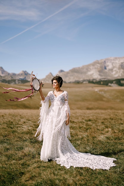 Bride in a white wicker dress with a tambourine in her hands stands in a field