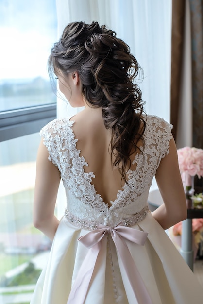 Bride with a beautiful hairstyle for long hair in a white dress stands back