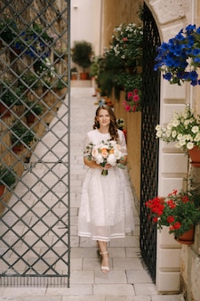 La sposa con un mazzo di fiori esce da un vecchio cortile con un cancello decorativo