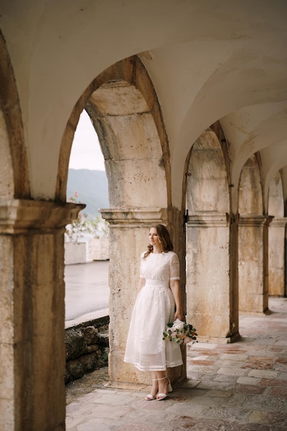 Bride with a bouquet of flowers stands in an ancient archway