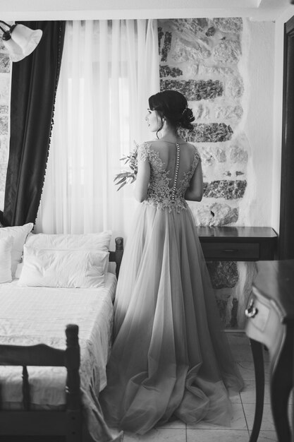 Bride with a bouquet of flowers stands near the bed in the room back view black and white photo