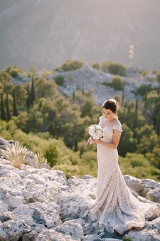 La sposa con un mazzo di fiori si erge sulle rocce sullo sfondo di montagne e vegetazione