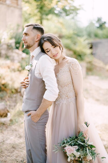 Bride with a bouquet in her hand hugs groom from behind with her head on his shoulder