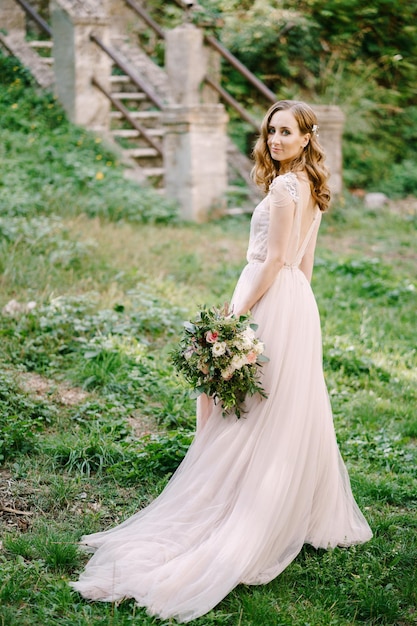 Bride with a bouquet stands halfturned on the green grass in front of a stone staircase