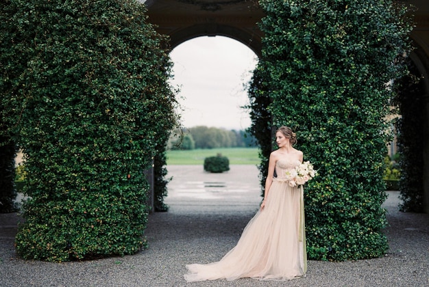 Bride with a bouquet stands near arches overgrown with green ivy Como Italy