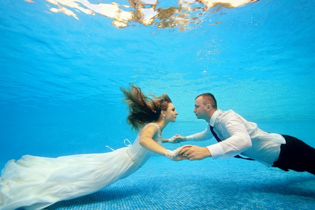 写真 乱れた髪の花嫁は花婿に向かって水中を泳ぎ、下の近くで彼の手を取ります