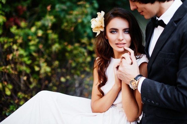 Bride with long curly hair and bridegroom standing close to each other at green leaves background, wedding photo.