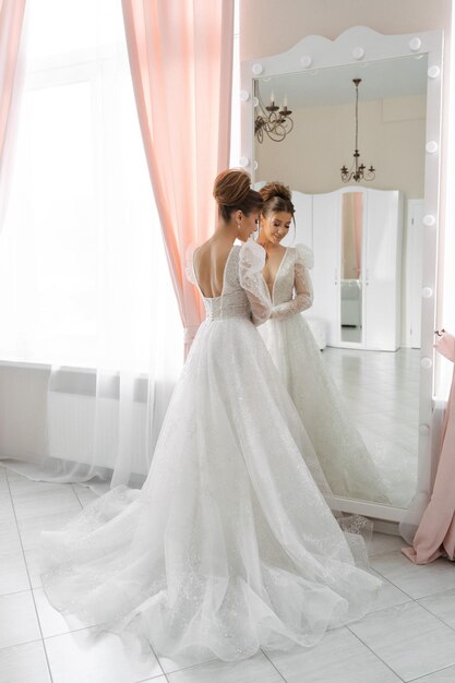 A bride with a train of a wedding dress stands at the mirror with her back to the camera in a bright interior