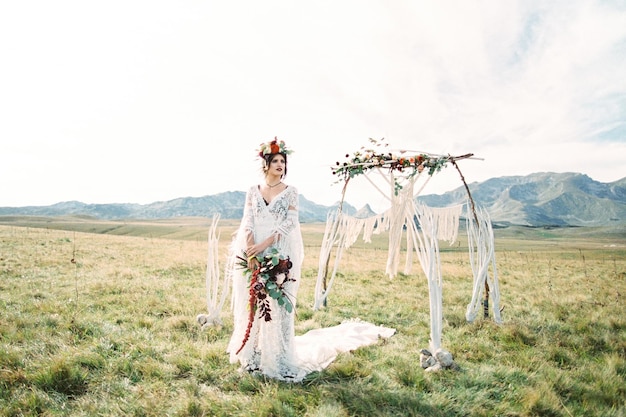 Bride in a wreath with a bouquet stands near a wedding arch in the lawn