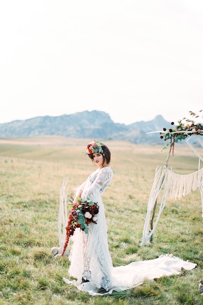 La sposa in una corona con un bouquet si erge su un tappeto bianco vicino a un arco nuziale