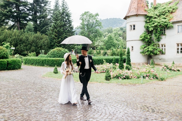 Photo brides in hats young girl in a white wedding dress and hat with a bouquet of flowers the bride and groom go and hold an umbrella brides on the background of the castle