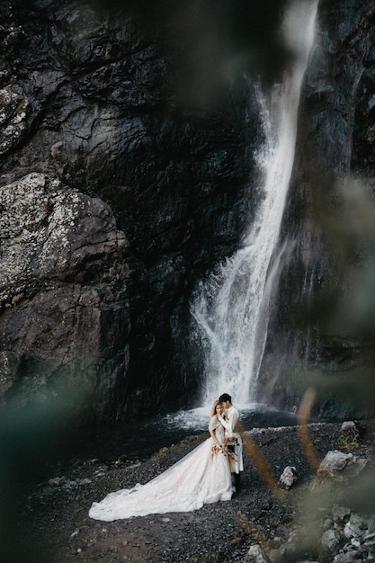 Spose che si abbracciano e posano per una foto sull'incredibile sfondo delle montagne rocciose con una cascata