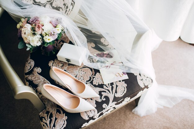 Brides wedding shoes with a bouquet with roses and other flowers on tha arm chair