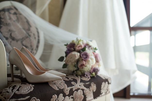 Brides wedding shoes with a bouquet with roses and other flowers on tha arm chair