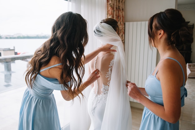 Photo bridesmaid helping bride fasten lacing her wedding white dress before ceremony.