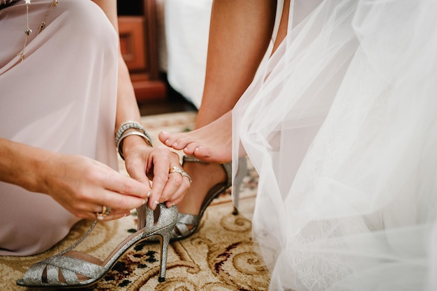 Bridesmaids put on leg bride stylish classic silver shoes Elegant Female putting hands on shoes on a background white dress Wedding morning preparation