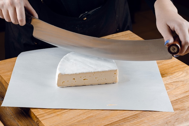 Brie soft white cheese from cow milk. Slicing brie on the wooden table. Organic delicious food.